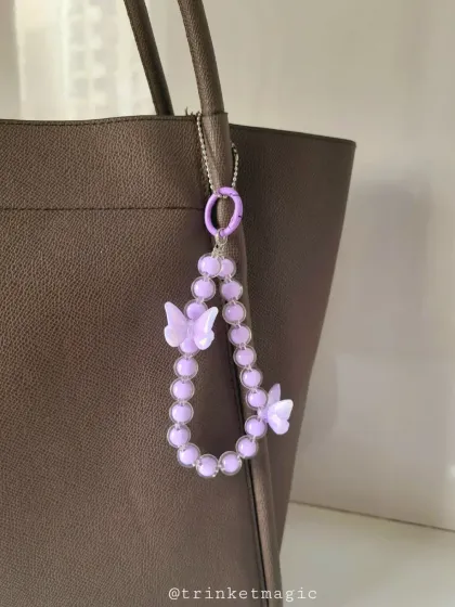 A simple yet elegant purple beaded bag charm with butterfly accents, shown on a dark tote bag. It's a great way to add a subtle pop of color and whimsy to your everyday carry.