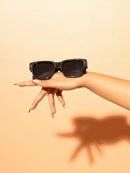 A hand with long nails playfully balancing a pair of black sunglasses. The clean, peach-colored background and sharp shadow create a modern, stylish look.