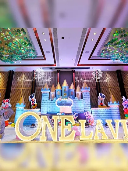The main stage of the Disneyland-themed party, featuring a large castle backdrop, marquee letters spelling "ONE LAVIN," and various Disney character cutouts. The green glass ceiling adds to the grand ambiance.