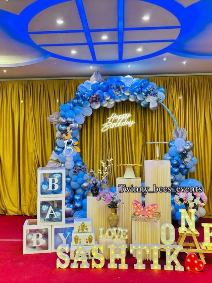 A classic Boss Baby theme for Sashtik's first birthday. The setup includes a blue and silver balloon arch, "BABY" blocks, and marquee letters spelling out his name against a gold curtain backdrop.