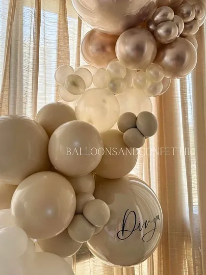 A close-up of the neutral-toned balloon garland, showing the mix of beige, cream, and rose gold chrome balloons. The personalized 'Divya' balloon adds a special touch.
