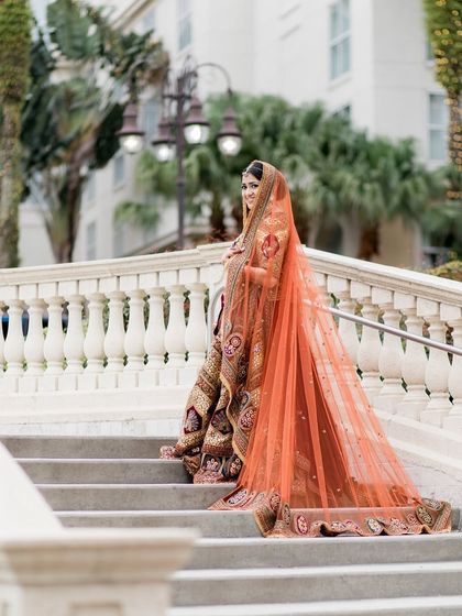 Ascending the stairs, the bride's long, orange-hued veil trails behind her, adding a touch of drama to her traditional, multi-toned lehenga.
