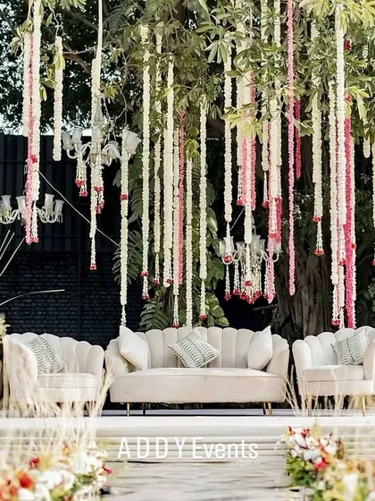 A serene mandap setup before the ceremony begins. The clean white seating, hanging floral garlands, and natural tree backdrop create a peaceful and elegant atmosphere for a day wedding.