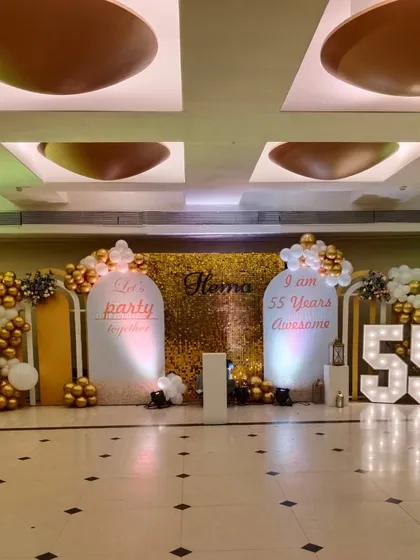 This image captures the complete 55th birthday stage setup. The lighting highlights the different textures of the gold shimmer wall, the smooth white panels, and the metallic shine of the balloons.