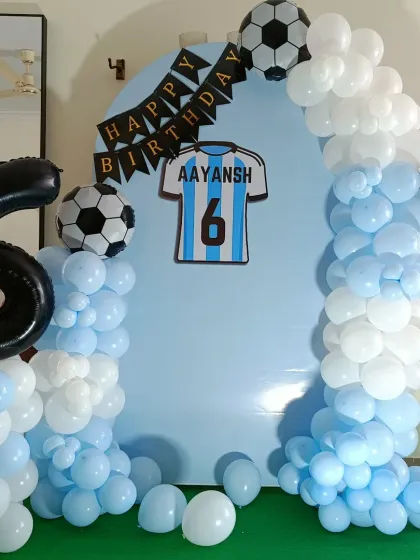A simple yet striking football-themed birthday setup. The light blue backdrop with a jersey graphic is framed by a white and blue balloon arch with football foil balloons.