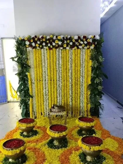 A traditional Haldi or Pooja setup with a backdrop of marigold and tuberose strings. The floor is decorated with a floral rangoli and brass bowls filled with rose petals.