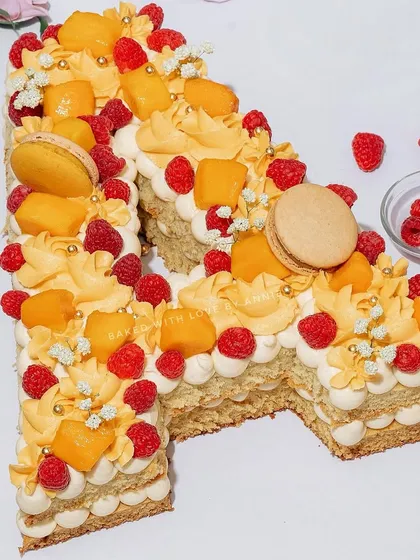 A custom monogram cake for a special celebration. This 'A' is made from layers of vanilla cake and cream, decorated with mango curd, fresh mango chunks, raspberries, and macarons.