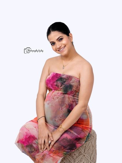 A happy and relaxed studio portrait of a mother-to-be in a colorful abstract dress, seated on a bohemian-style stool.