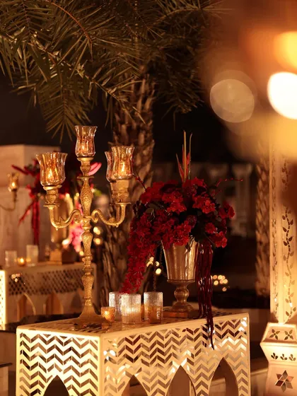A detail from a night event, where a golden candelabra and a vase of red flowers are set against an illuminated jali console. The warm, glowing light creates a magical and intimate atmosphere.