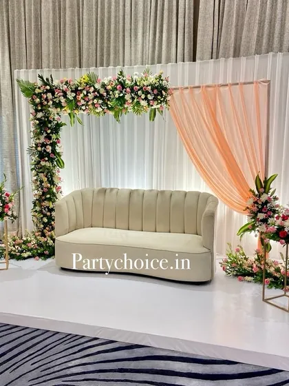 A simple yet elegant reception stage setup. The decor features a modern sofa on a white platform, framed by an L-shaped floral arch and accented with a single peach-colored drape for a touch of softness.
