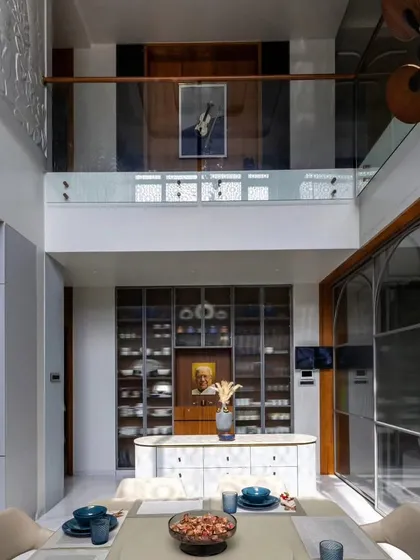 This view showcases the grandeur of a double-height dining and living area we designed. The glass-railed upper gallery creates a visual connection between the two floors, enhancing the sense of space. We installed a custom glass-fronted crockery unit and an elegant island counter to define the dining space within the open-plan layout.