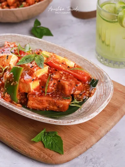 A plate of saucy chili paneer, looking glossy and delicious. The fresh basil garnish and refreshing drink in the background complete the meal setting.