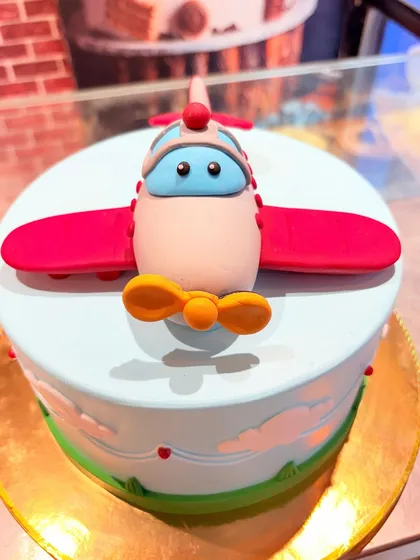 A cute airplane-themed cake with a friendly-faced plane made of fondant. The cake is decorated with clouds and a green landscape.