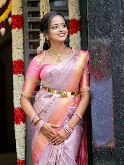 A smiling bride in a lavender Kanchivaram with a pink border. The matching pink blouse has simple gold detailing, creating a classic and elegant look.