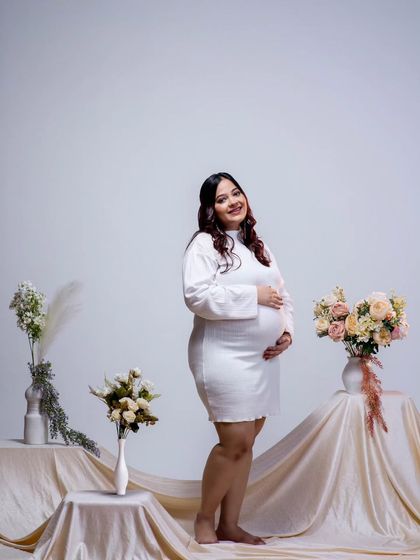A full-length solo portrait showcasing the beauty of the final trimester. The simple white dress and clean background keep the focus on her radiant smile and the baby bump.
