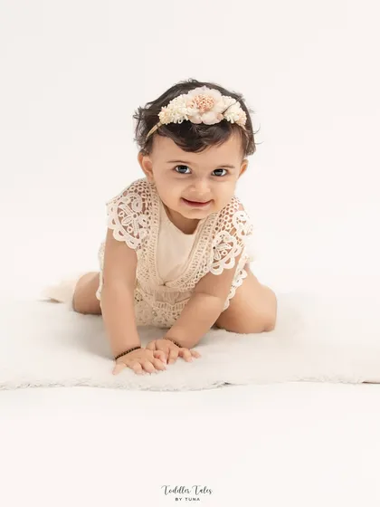 A baby girl crawls on a soft white rug, looking up with a bright, happy smile. This is a classic, simple portrait from a family session, focusing on the child's joy.