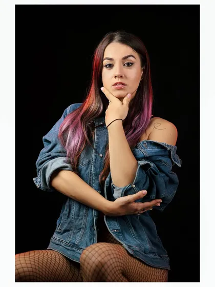 A casual studio portrait of a model with colorful hair, wearing a denim jacket and fishnet stockings against a black background.