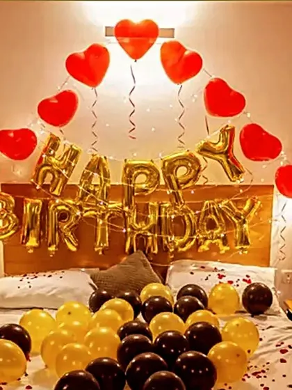A classic birthday room decoration. The bed is decorated with yellow and black balloons, while the wall features 'Happy Birthday' foil letters, heart balloons, and champagne-themed props.