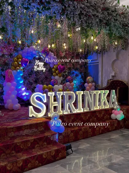 I can transform any stage into a fairytale setting. This first birthday stage decoration features large illuminated letters for the name, a 'Happy Birthday' neon sign, and a stunning backdrop of flowers and lights, creating a dreamy atmosphere.