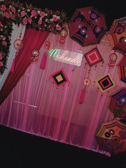 A full view of a Mehendi stage setup. The backdrop features a pink floral arch, a 'Mehendi' neon sign, and a wall decorated with colourful umbrellas and traditional hangings, all set on an artificial grass floor.
