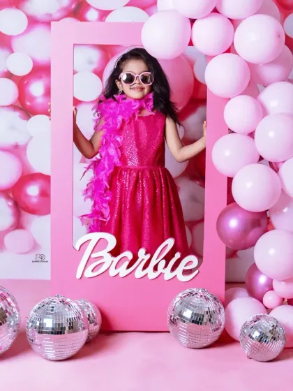 A vibrant 'Barbie' themed photoshoot for a stylish toddler. She poses confidently in a pink frame against a wall of pink balloons and disco balls, complete with a feather boa and sunglasses.