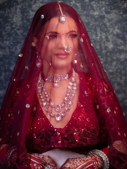 A beautiful shot of a bride under a sheer red veil. The makeup is soft and romantic, with a focus on glowing skin and defined eyes that look mesmerizing even through the dupatta.