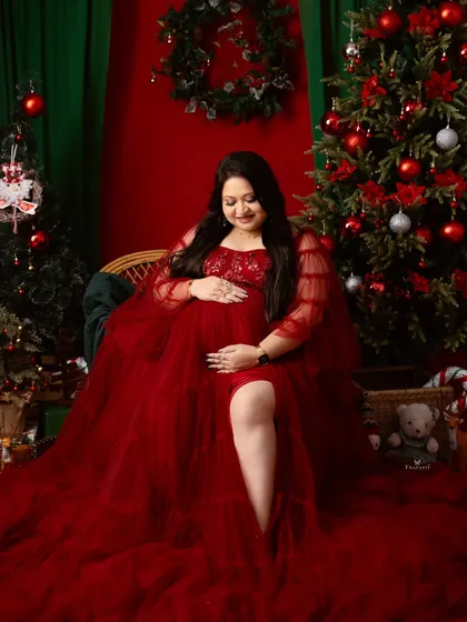 A festive and glamorous portrait from a Christmas-themed shoot. The mom-to-be is seated amidst holiday decorations, her red gown adding to the celebratory feel.