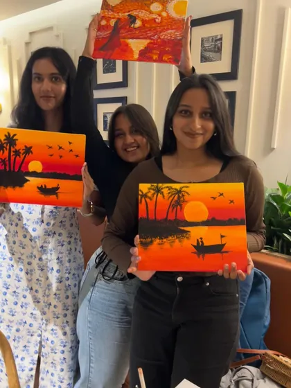 Three friends holding up their sunset paintings, with one playfully holding her art above her head.