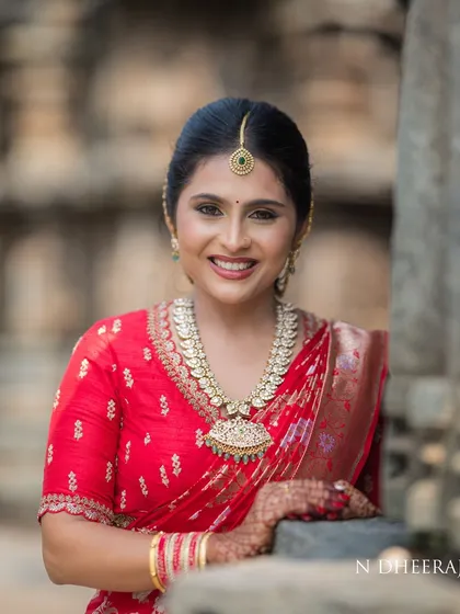A close-up portrait of a bride in her red saree. The intricate neckline embroidery on her blouse is visible, complementing her traditional coin necklace.