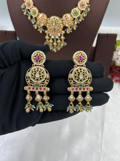 This antique gold necklace set is crafted with intricate filigree work and studded with ruby and emerald-colored stones. The matching earrings complete this traditional and elegant look.