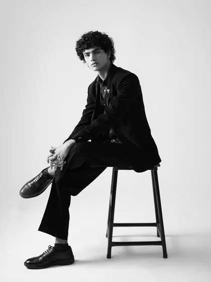 A black and white full-length portrait for a menswear lookbook. The classic seated pose is sophisticated and provides a clear view of the suit's tailoring and fit.