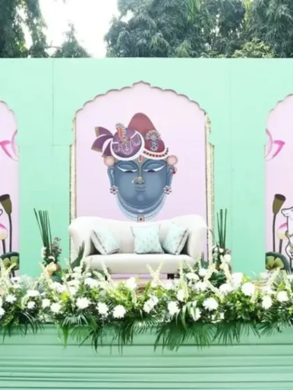 This Mehendi stage is designed around a Pichwai theme, featuring a custom backdrop with a traditional painting of Shrinathji. The seating area is framed with fresh white flowers and set against a pastel green structure.