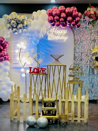 Here is a beautiful birthday decoration featuring a half-arch of pink, silver, and white balloons against a shimmering sequin wall. The "Happy Birthday" neon sign and elegant props like the decorative fence and cake stands complete this chic and modern look.
