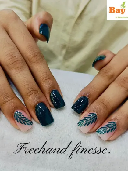 Freehand finesse on display. This design features short, square nails with a deep teal polish and two accent nails with a nude base and intricate green leaf patterns.