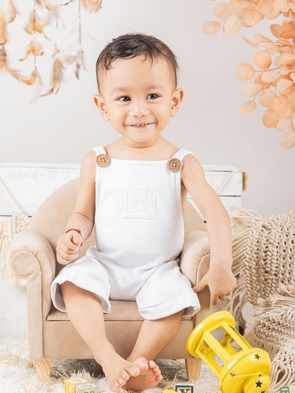 A classic portrait of a baby boy in a white romper, sitting in a tiny armchair.