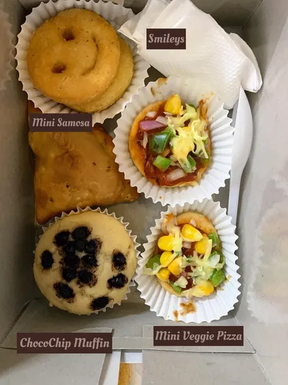 A snack box designed to bring smiles, especially for kids' parties. It includes a Choco-Chip Muffin, Mini Veggie Pizza, a Mini Samosa, and fun potato Smileys.