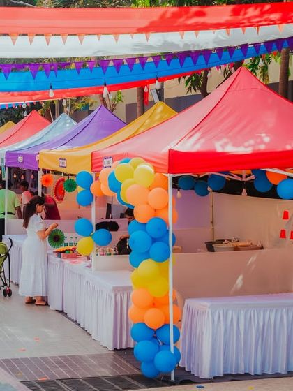 A wide shot of the colorful flea market stalls at the CNS Fiesta. The vibrant canopies and balloon decorations create a lively and inviting shopping experience.