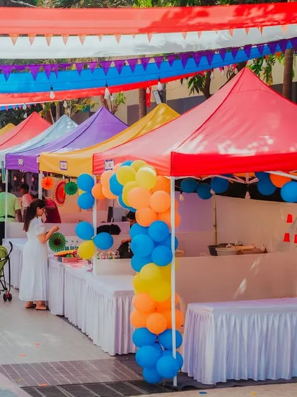 A wide shot of the colorful flea market stalls at the CNS Fiesta. The vibrant canopies and balloon decorations create a lively and inviting shopping experience.