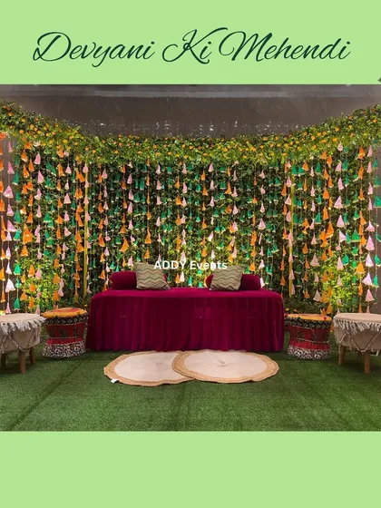 A vibrant Mehndi stage with a backdrop of green foliage and colorful hanging tassels, creating a festive and playful setting.