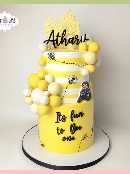 It's fun to bee one! This cheerful two-tiered cake for a first birthday has a bee theme, with yellow and white stripes, cute edible bees, and honeycomb details.