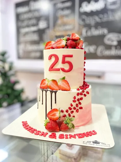 A two-tiered 25th-anniversary cake featuring fresh strawberries, pomegranate seeds, and a chocolate drip.