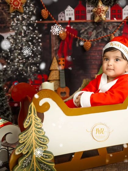 A classic Christmas portrait of a baby in a Santa suit, sitting in a sleigh surrounded by festive decorations.