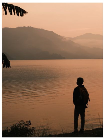 A traveler stands silhouetted against the golden light of sunset over Phewa Lake, a moment of quiet contemplation.