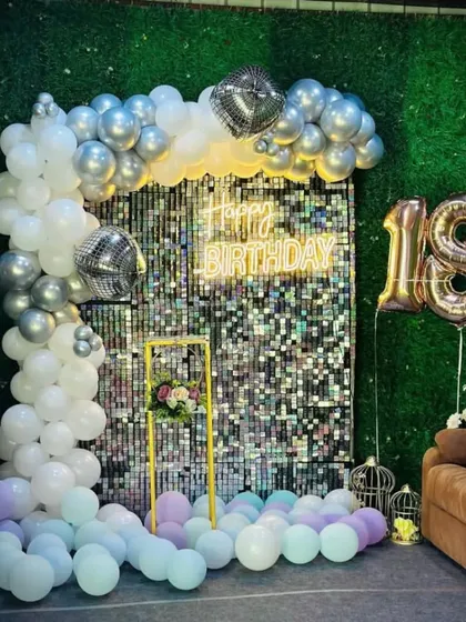 An 18th birthday celebration with a silver sequin wall and a green hedge backdrop. The decoration features a white and silver balloon arch and gold foil number balloons.