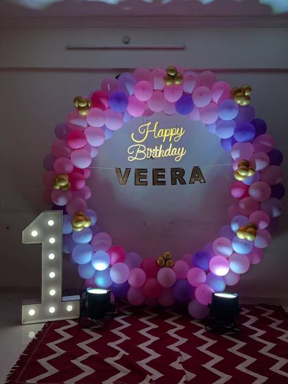 A circular balloon frame in shades of pink and purple for Veera's first birthday. The setup is completed with a "Happy Birthday" sign and a large light-up number '1'.