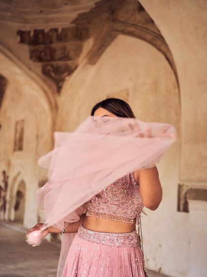 An artistic shot where the dupatta flows in front of the model, creating a sense of mystery and movement. The focus is on the texture and color of the pink lehenga.