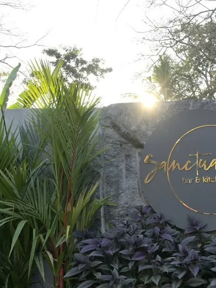 The entrance to Sanctuary Bar & Kitchen, with its name elegantly displayed on a stone wall. The surrounding tropical plants and purple foliage create a vibrant and welcoming first impression.