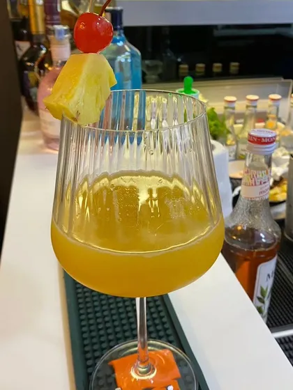A close-up of a vibrant yellow cocktail in a ribbed wine glass, garnished with pineapple. Our use of fresh fruit and quality glassware elevates the drinking experience.