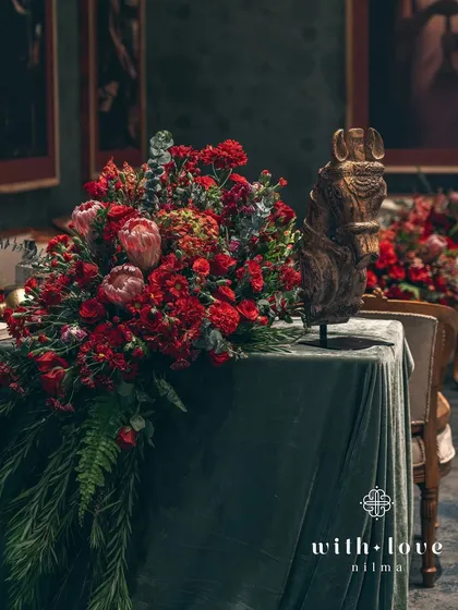 A dramatic floral arrangement cascading over a velvet-draped table. The composition is bold and artistic, reflecting the spirit of Raja Ravi Varma.