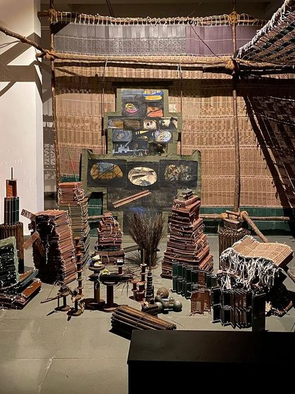 An installation view of 'WEAVING LIGHT', showing stacks of textile-related objects and a large loom-like structure. The exhibition is a testament to the evolution of handcrafted textiles.